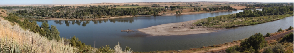 [Four photos stitched together showing the river with an island in the middle thus making it appear the river is split into two streams. A lot of sand is visible at one end of the island. Trees line the edges of the river.]